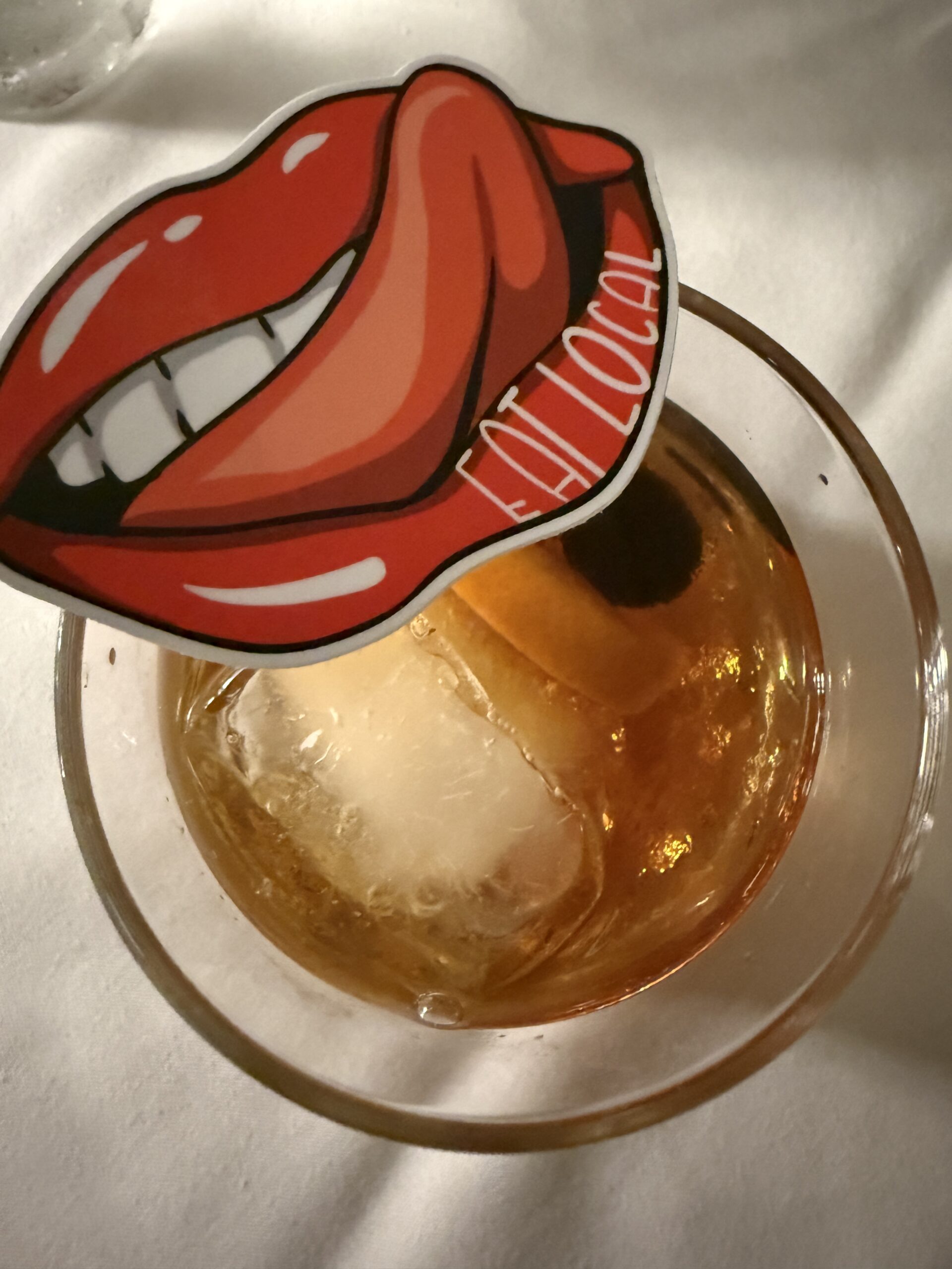 old-fashioned cocktail with a sticker of red lips and a tongue with "eat local" printed on it"